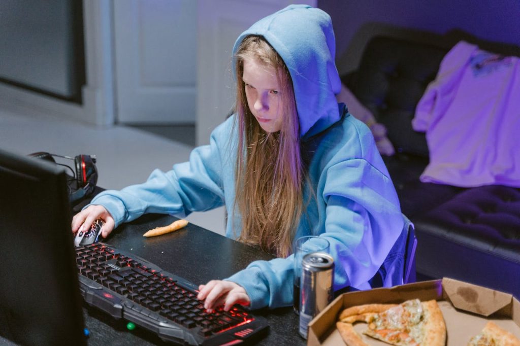 Teenage girl in blue hoodie gaming indoors with pizza and energy drink nearby.
